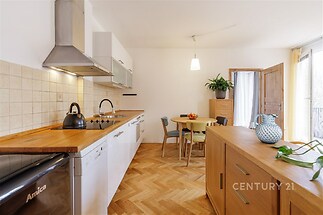 Světlá kuchyň s parketovou podlahou, moderní spotřebiče, dřevěná pracovní deska, velká okna poskytující dostatek přirozeného světla.