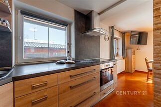 Moderní kuchyň s dřevěnou kuchyňskou linkou, vestavěnými spotřebiči a dlažbou, velká okna zajišťují světlou a přívětivou atmosféru.