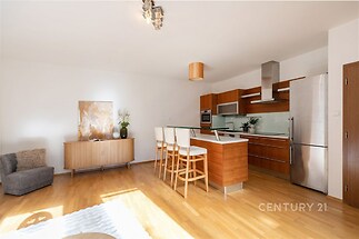 Moderní kuchyň s parketovou podlahou, ostrůvkem a vestavěnými spotřebiči. Velká okna zajišťují dostatek světla a dodávají prostoru svěží dojem.