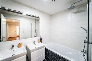 Moderní koupelna s bílými obklady, vanou a sprchou, dvojitým umyvadlem a vestavěným osvětlením, světlý interiér.
