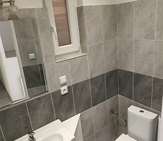 Malá koupelna s moderní dlažbou v šedém tónu, keramické umyvadlo s bílou skříňkou a okno pro přirozené osvětlení.