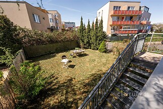 Zahrada s travnatou plochou a zahradním nábytkem, obklopená keři a stromy, nabízí soukromí. Moderní bytové domy v pozadí zvýrazňují atraktivitu místa.