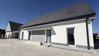 Moderní rodinný dům s bílou fasádou, sedlovou střechou a velkými okny, umístěný na vydlážděném pozemku pod modrou oblohou.