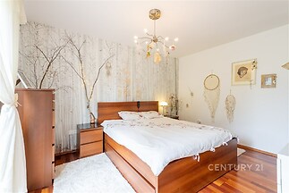 Prostorná ložnice s parketovou podlahou, světlými stěnami a moderním osvětlením, dřevěný nábytek, dekorativní tapeta s přírodním motivem.