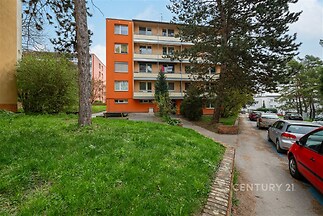 Čtyřpodlažní bytový dům s oranžovou fasádou a balkony, obklopený zelení a vzrostlými stromy, nacházející se v klidné rezidenční čtvrti.