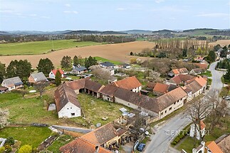 Pohled na venkovský statek s červenými střechami, obklopený zelenými poli a nízkými domy, ideální pro rekonstrukci nebo investici.