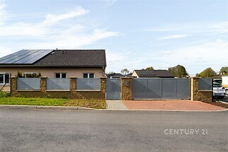 Venkovní pohled na moderní rodinný dům se solárními panely, zděný plot s kovovými prvky, udržovaná zahrada, asfaltová příjezdová cesta.