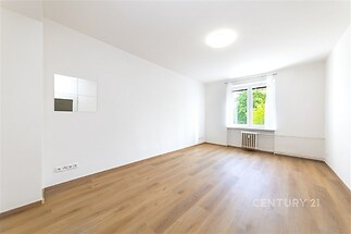 Světlý obývací pokoj s parketovou podlahou, velká okna nabízejí přírodní světlo, moderní styl a čistá bílá výmalba zdi.