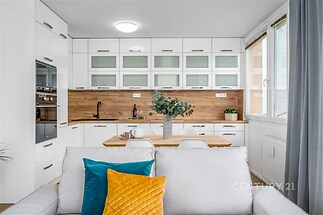 Moderní kuchyň s bílou vestavěnou linkou, dřevěnou pracovní deskou a parketovou podlahou, velká okna propouštějí dostatek světla.