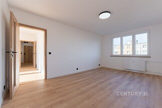Světlý obývací pokoj s dřevěnou podlahou, velkými okny a moderním osvětlením, ideální pro útulné bydlení v rekonstruovaném bytě.