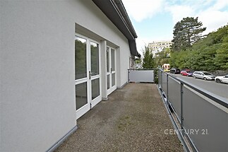 Balkón s kovovým zábradlím, na zemi betonový povrch, moderní styl, velká francouzská okna do obývacího pokoje, pohled na klidnou ulici a zeleň.