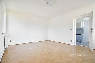 Světlý obývací pokoj s parketovou podlahou, velkými okny a světlými stěnami vedoucí do moderní kuchyně v rekonstruovaném bytě.