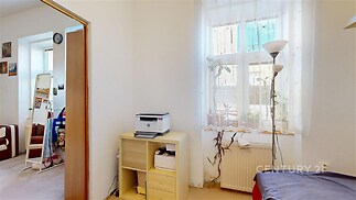 Světlá ložnice s velkým oknem, parketová podlaha a moderní zařízení, samostatná police a pokojové rostliny vytvářejí útulný prostor.