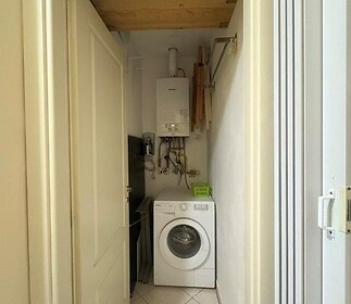 Úzká prádelna s dlažbou, obsahuje pračku a plynový kotel. Praktický prostor s úložnými policemi a světlým designem.