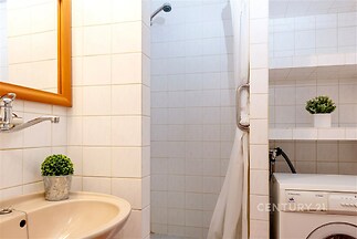 Retro koupelna s bílými kachličkami, sprchovým koutem, umyvadlem a pračkou pod policí, původní stav.