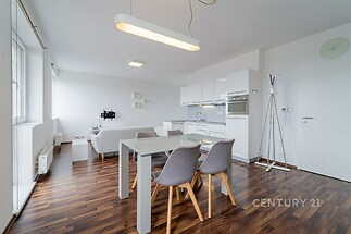 Moderní obývací pokoj s kuchyňským koutem, parketová podlaha, bílá kuchyňská linka, světlý interiér, velká okna pro více světla.
