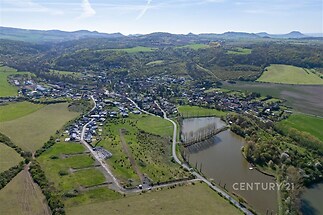Satelitní pohled na malebnou vesnici obklopenou zelenými poli a lesy, s rybníkem v popředí a horami na horizontu.