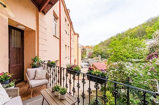 Balkon s dřevěnou dlažbou, výhledem na zelené prostředí a ozdobným kovovým zábradlím, vhodný pro relaxaci v klidné části města