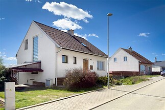 Rodinný dům s moderní fasádou, šikmou střechou a velkými okny, nacházející se na klidné ulici s dlážděným chodníkem.