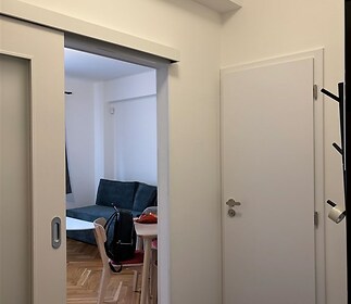 Vstupní předsíň s parketovou podlahou a moderními posuvnými dveřmi, vedoucí do světlého obývacího pokoje s minimalistickým nábytkem.