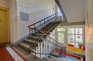 Světlé schodiště s dlažbou, velkými okny a původním zábradlím, vhodné pro bytový dům, zachovalý historický styl.