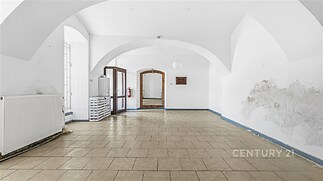 Prostorný pokoj s dlažbou, velkými okny a klenutým stropem, ideální pro rekonstrukci, nabízí různé možnosti využití díky své velikosti a světlu.