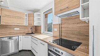 Moderní kuchyň s dřevěnými dvířky, vestavěnými spotřebiči a dlažbou. Velká okna zajišťují světlé prostředí.