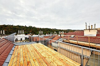 Střešní terasa s dřevěnou podlahou, nabízející výhled na okolní střechy a zeleň, vhodná pro relaxaci a venkovní posezení.