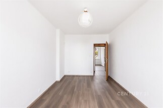 Prázdný pokoj s dřevěnou podlahou, bílými stěnami a otevřenými dveřmi. Velká okna zajišťují světlé prostředí.