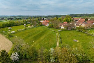 Letecký pohled na malebnou vesnici obklopenou zelenými loukami a cestou; ideální místo pro klidné bydlení v prostředí přírody.