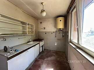 Zchátralá kuchyň s velkými okny, původními skříňkami a starými dlaždicemi, ideální k rekonstrukci a modernizaci.