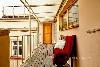 Zastřešená terasa s dřevěnou podlahou, moderní zábradlí a velká okna poskytující dostatek světla. Pozornost přitahuje pohodlná lavička.