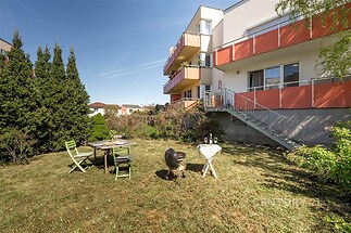 Zahrada u moderního bytu s terasou, posezením a grilem, ideální pro venkovní relaxaci a společenské aktivity, upravený trávník a vzrostlé keře.