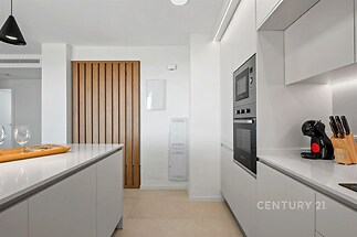 Moderní kuchyň s bílou linkou, vestavěnými spotřebiči a dřevěnými prvky. Dlažba na podlaze dodává prostoru elegantní vzhled.