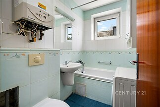 Koupelna s dlažbou a vanou, světlá díky velkým oknům, obsahuje plynový kotel a pračku, vhodná k renovaci