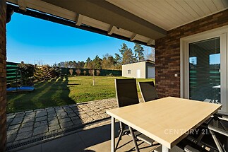 Krytá terasa s dlažbou, přes kterou je výhled na prostornou zahradu s rovnou trávníkovou plochou a dřevěným plotem, nabízí klidné prostředí.