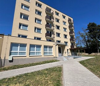 Vícepodlažní panelový bytový dům, zrenovovaný fasádou, s balkony a přístupovým chodníkem, obklopený zelení, ideální pro rodiny