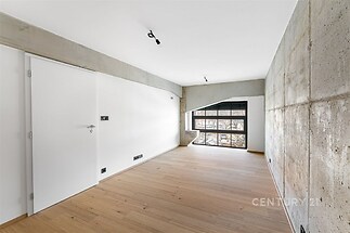 Světlý obývací pokoj s dřevěnou parketovou podlahou a velkými okny, moderní design s betonovou stěnou.