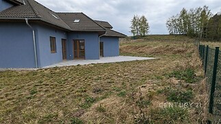 Rodinný dům s šedou fasádou, terasou a zahradou. Moderní stavba s velkými okny a střešními okny. Přírodní okolí nabízí klid a soukromí.