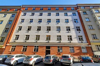 Čtyřpatrová cihlová fasáda bytového domu s mnoha okny, nacházející se na rušné městské ulici, dobře udržovaná, s parkováním před domem.