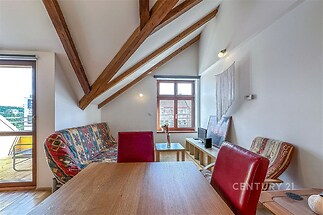 Podkrovní obývací pokoj s dřevěnými trámy, velká okna a stylový interiér, který zahrnuje světlý dřevěný nábytek a barevné sezení.