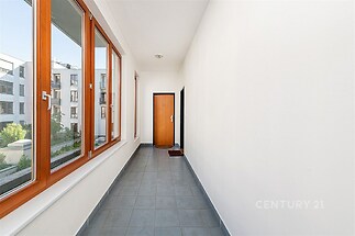 Dlouhá světlá chodba s velkými okny, dlažbou a moderním designem, poskytující výhled do okolí a přístup do sousedních bytů