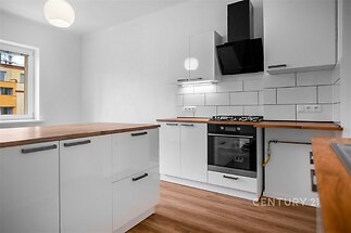 Moderní kuchyň s dřevěnou podlahou, bílými skříňkami a vestavěnou troubou, světlý prostor s velkým oknem a moderním digestořem.