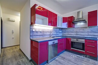 Moderní kuchyň s parketovou podlahou, červenými lesklými skříňkami a vestavěnými spotřebiči, včetně myčky a trouby, nabízí svěží design.