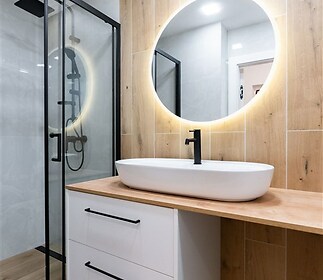 Moderní koupelna s dřevěnými obklady, skleněným sprchovým koutem a kulatým osvětleným zrcadlem vytváří elegantní vzhled.