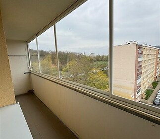 Zasklená lodžie s dlažbou, výhledem na zeleň a okolní panelové domy, vhodná pro relaxaci nebo pěstování rostlin.