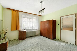 Prázdný pokoj s kobercem, dřevěná šatní skříň a starožitné vybavení, světlá záclona, místnost připravená k rekonstrukci nebo modernizaci.