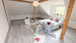 Světlá ložnice s dřevěnými trámy, laminátovou podlahou a velkým oknem nabízí moderní styl a útulnou atmosféru.