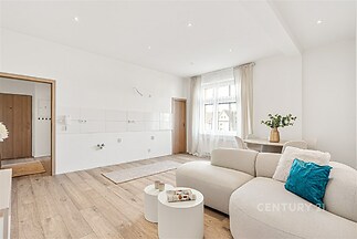 Světlý obývací pokoj s parketovou podlahou, velkými okny a moderním nábytkem. Prostor nabízí rekonstrukci a otevřený koncept s přírodním osvětlením.