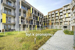 Moderní bytový komplex s balkonem, velká okna a upravený travnatý prostor. Stavba je moderní s pestrobarevnými fasádami.